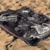 Aquarius Flow woven blanket with skeleton hand and water design lying on sandy surface.