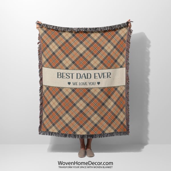 Woman holding up the fully unfolded "Best Dad Ever" blanket with a plaid pattern in warm orange and beige tones; her face is not visible.