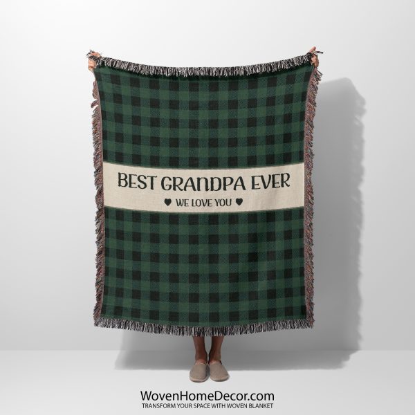 Woman holding up the fully unfolded "Best Grandpa Ever" blanket with a green plaid pattern; her face is not visible.