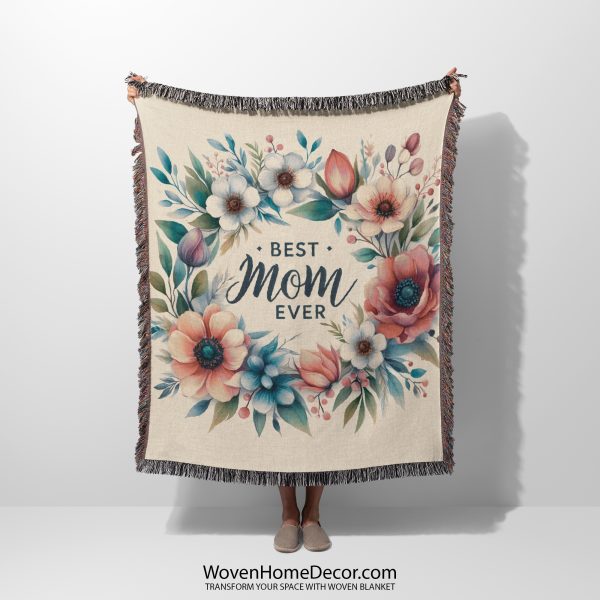 Woman holding up the fully unfolded "Best Mom Ever Bloom Woven Blanket" with a floral wreath of daisies and pastel blooms surrounding the text “Best Mom Ever”; her face is not visible.