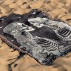 Gemini Duality woven blanket with twin skeletons lying on sandy surface.