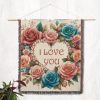 The "Love Blooms Woven Blanket" hanging on a light-colored wall as a tapestry, displaying its intricate floral wreath and loving message.