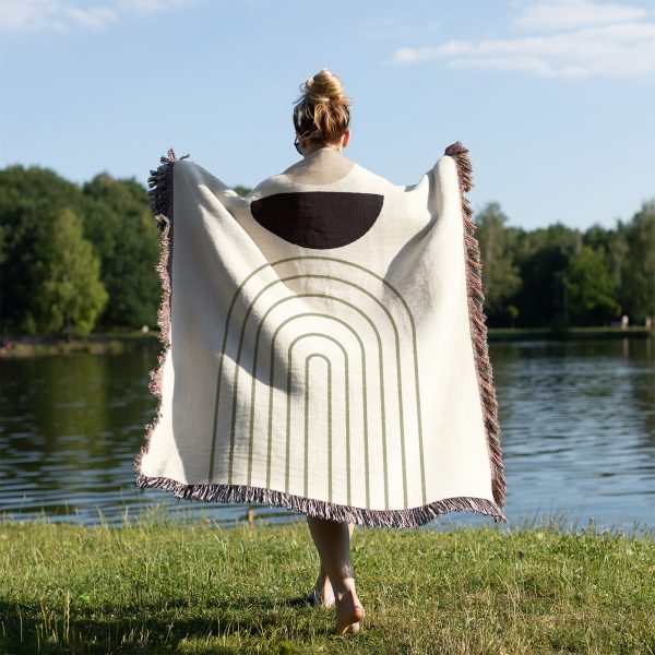 Person standing near a lake, seen from the back, wrapped in the Modern Arc Woven Blanket.