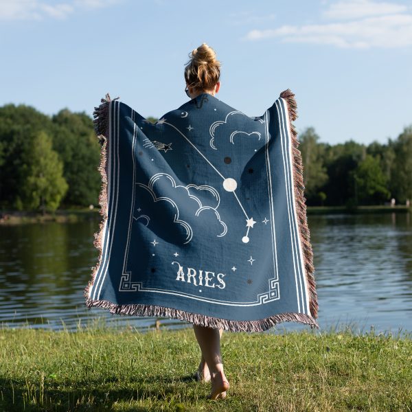 A woman stands by a peaceful lake, wrapping herself in a warm woven blanket featuring the bold Aries constellation, visible from behind.