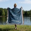 A woman standing by the lake, wrapped in a Capricorn zodiac woven blanket, with earthy tones and the mountain goat constellation design visible from behind.