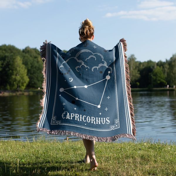 A woman standing by the lake, wrapped in a Capricorn zodiac woven blanket, with earthy tones and the mountain goat constellation design visible from behind.