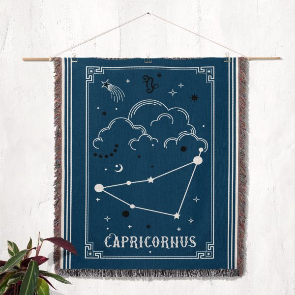 The Capricorn zodiac woven blanket hanging as a tapestry on the wall, showcasing the mountain goat constellation in earthy brown and green tones.