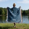 A woman standing near a lake, wrapped in a Libra zodiac woven blanket, with the design of balance scales visible as she enjoys the calm surroundings.