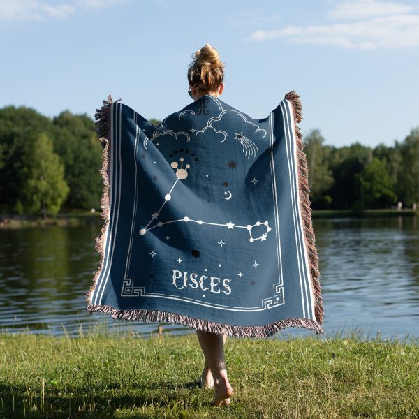 A woman standing by a tranquil lake, wrapped in a Pisces zodiac woven blanket, with the twin fish constellation design visible as she enjoys the peaceful surroundings.