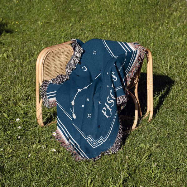 The Pisces zodiac woven blanket resting on a chair in the grass, with its soft blue and turquoise colors blending beautifully with the natural outdoor setting.