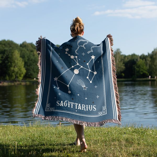 A woman stands near a lake, draped in a Sagittarius zodiac woven blanket, with the archer constellation design visible as she enjoys the outdoors.