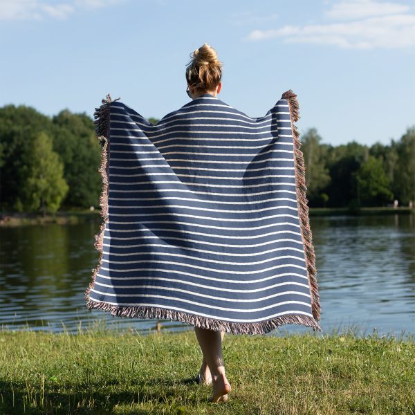 A person standing on grass near a lake, wrapped in the Scandinavian Blue Striped Woven Blanket.