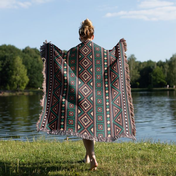 A person wrapped in an African-patterned woven blanket near a lake on grass.