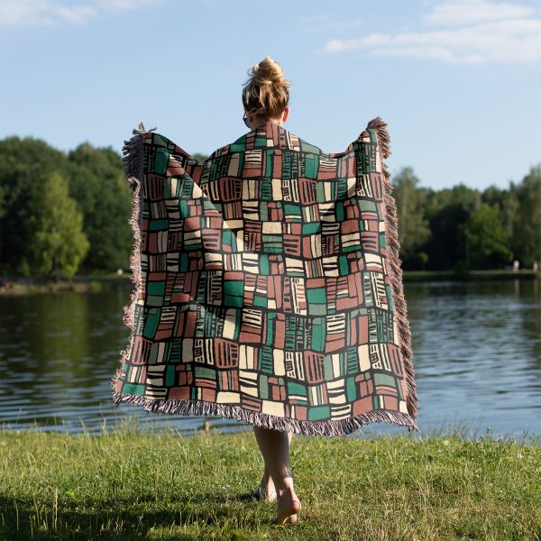 A person wrapped in a geometric-patterned woven blanket stands by a lake.