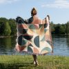 Woman standing with her back to the camera, covering herself with the modern geometric blanket on grass near a lake.