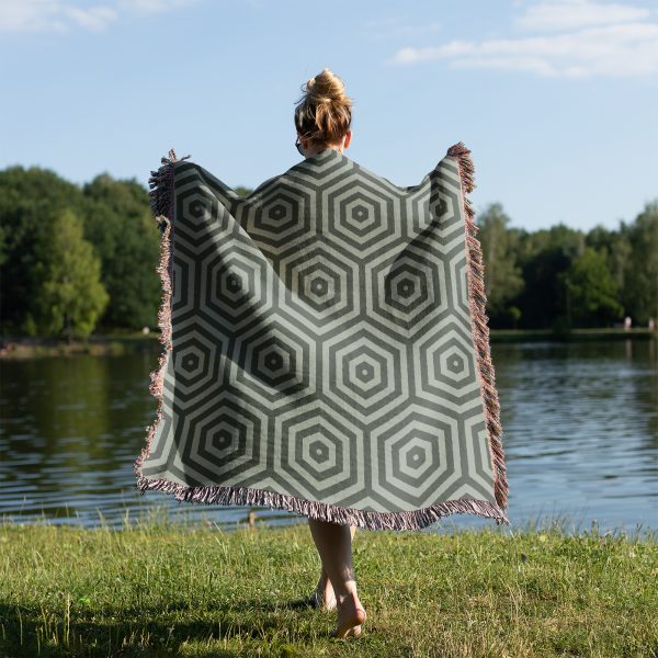 Woman standing with her back to the camera, covering herself with the woven blanket on grass near a lake.