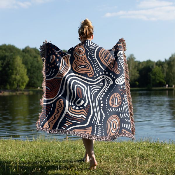 A person wrapped in an organic-patterned woven blanket stands near a lake on grass.