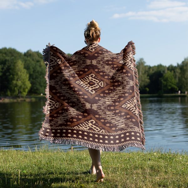 A person wrapped in a tribal-patterned woven blanket stands by a lake on grass.