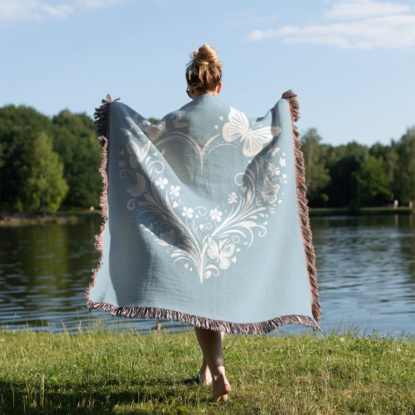 A person standing on a grassy lakeside, viewed from behind, draped with the woven blanket.