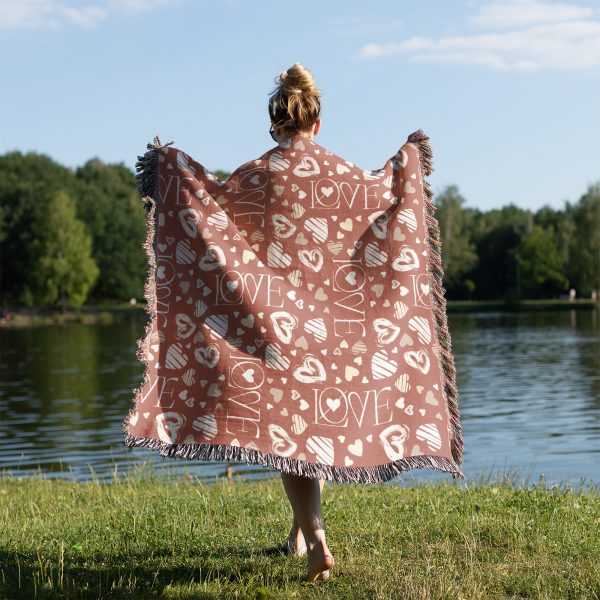 A woman stands by the lake, draped in the Love Symbols Woven Blanket, with her back facing the viewer.